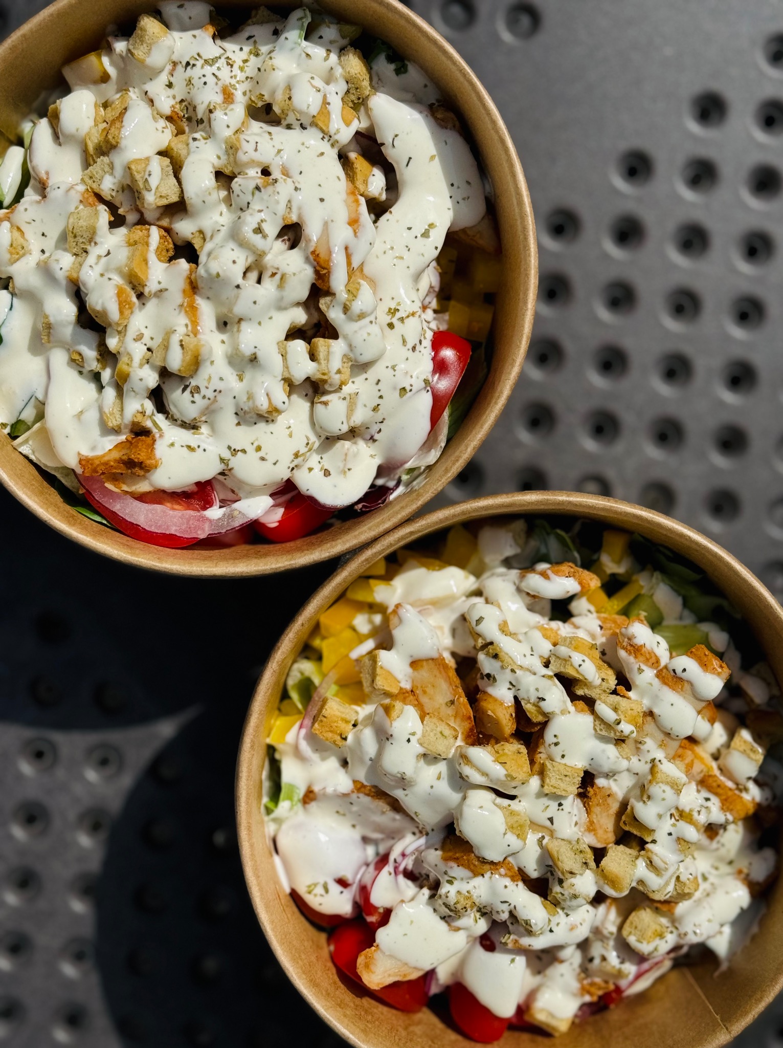 Dwa papierowe kubki z sałatką: grillowany kurczak, warzywa i sos. Całość posypana grzankami i ziołami. Widok z góry, ostre światło.