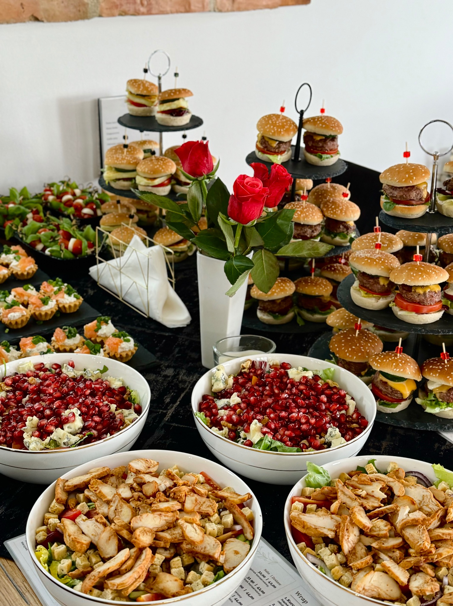 Elegancki stół cateringowy z mini burgerami, sałatkami z granatem i kurczakiem, tartaletkami z łososiem i bukietem czerwonych róż.