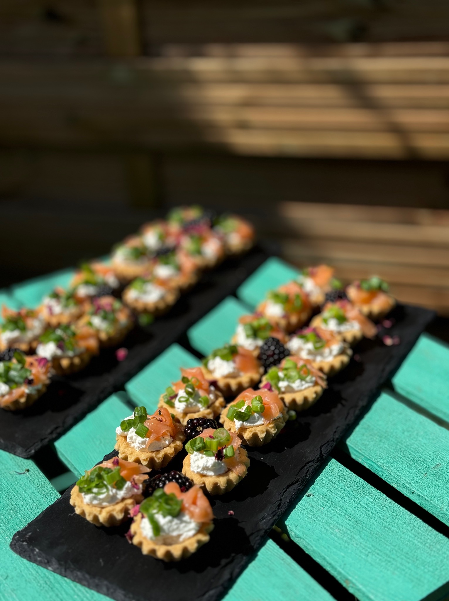 Dwie czarne deski z tartaletkami z łososiem, kremem i zielonym szczypiorkiem na turkusowym drewnianym stole w plenerze, w słoneczny dzień.