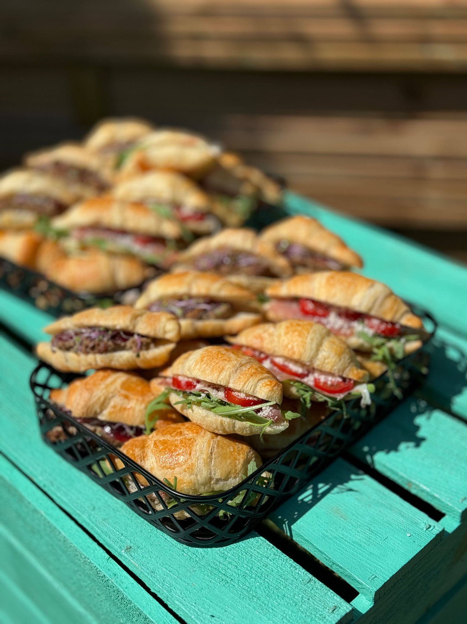 Kruche rogaliki z różnymi nadzieniami, ułożone w koszykach na turkusowym, drewnianym stole w słoneczny dzień. Catering na imprezę firmową.