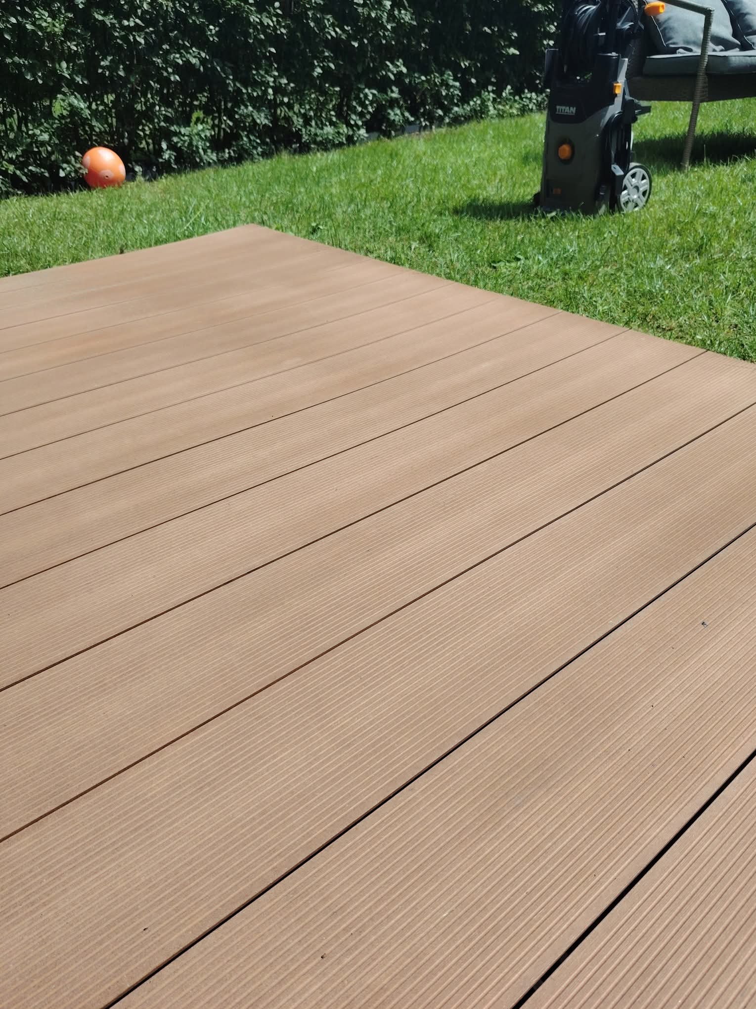 Czysty, brązowy taras kompozytowy po umyciu myjką ciśnieniową Titan, na tle zielonego trawnika i krzewów. Widoczny fragment sofy ogrodowej.