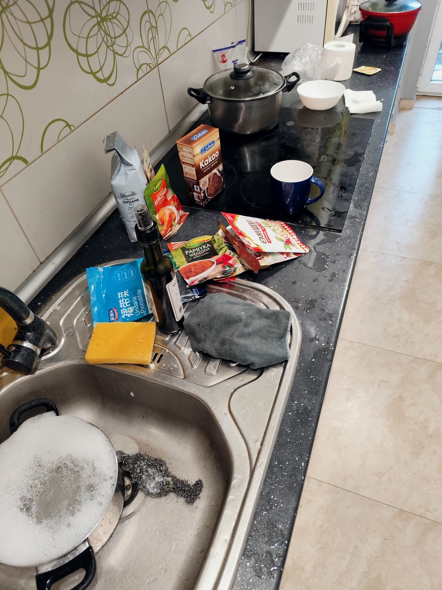 Zlew kuchenny ze stertą naczyń i akcesoriami do mycia, obok przyprawy, kakao i płatki owsiane. Na płycie resztki po gotowaniu. Widok na blat kuchenny z bliska.