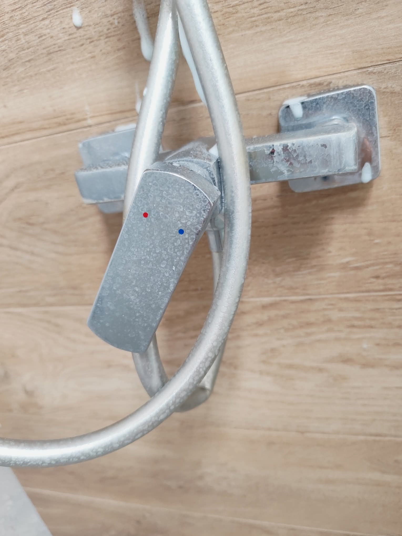 Z bliska: bateria prysznicowa z osadem kamiennym na tle drewnopodobnej ściany. Widoczne ślady zaschniętego mydła i krople wody.