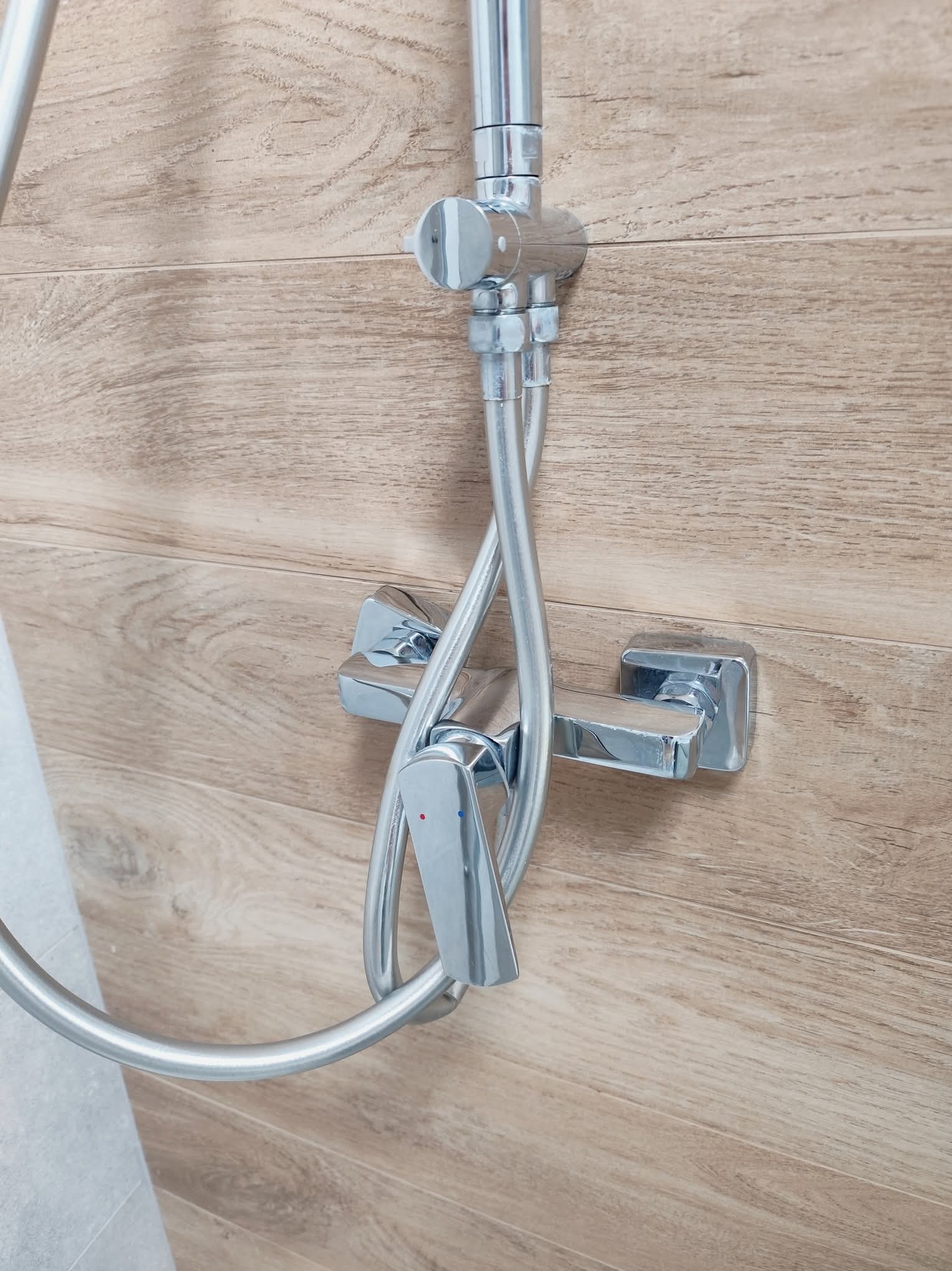 Błyszcząca, chromowana bateria prysznicowa zamontowana na ścianie wyłożonej panelami imitującymi drewno. Widoczny fragment węża prysznicowego i płytki podłogowej.