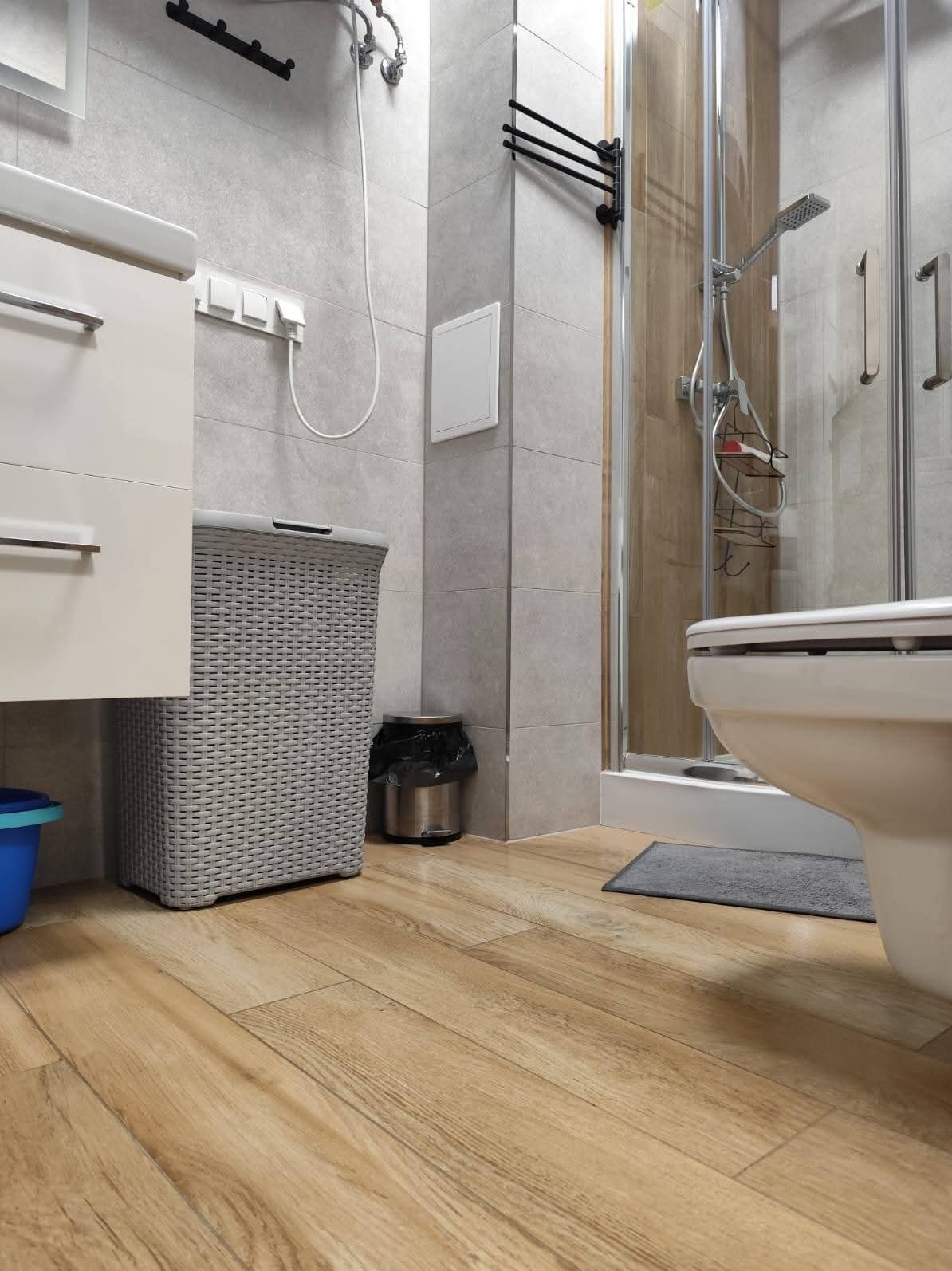 Jasna łazienka z drewnopodobną podłogą, szarą koszem na pranie i kabiną prysznicową. Widoczny fragment toalety i kosz na śmieci. Czyste, minimalistyczne wnętrze.