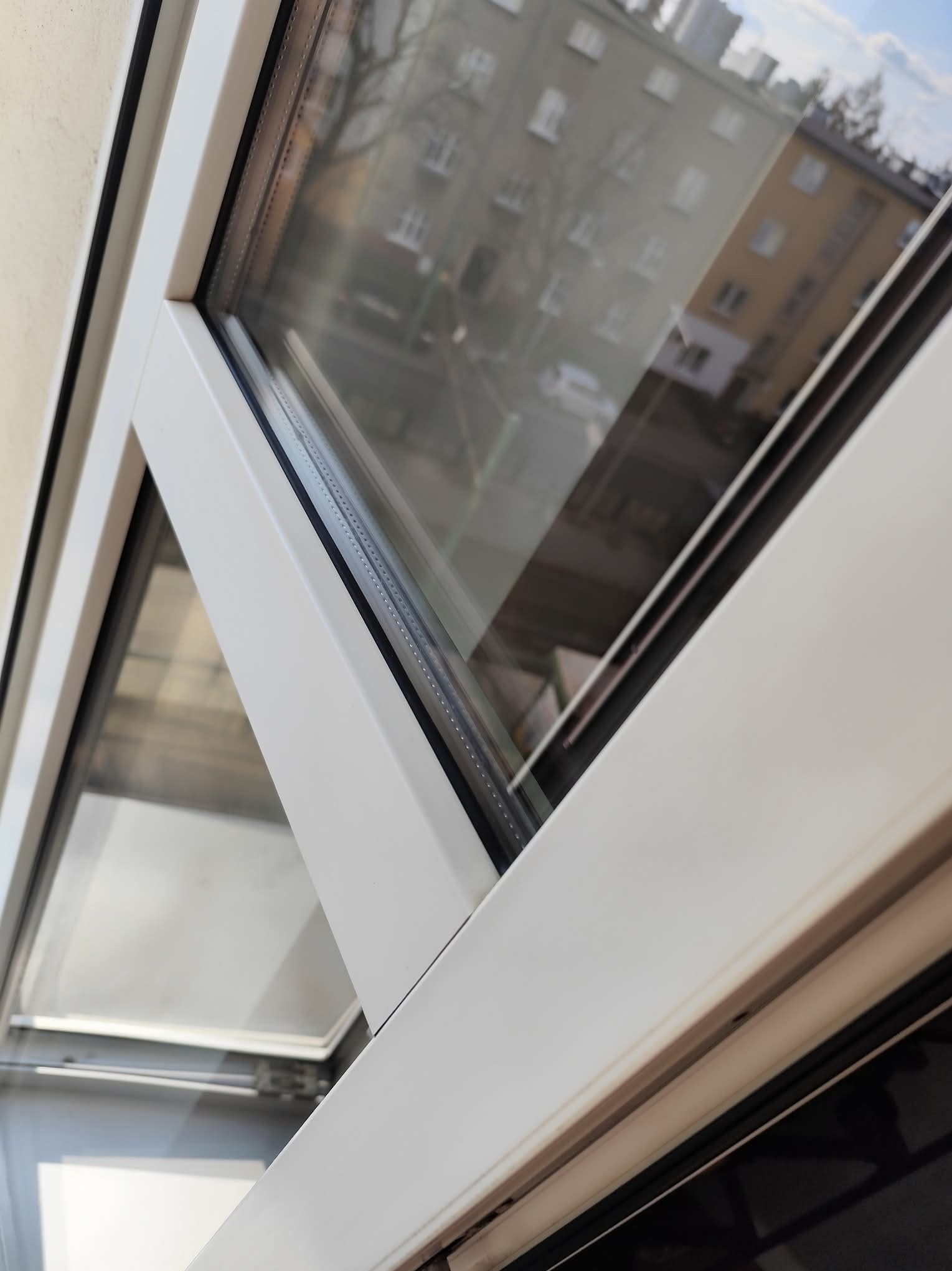 Białe, otwarte okno PCV z widokiem na budynki odbijające się w szybie. Widoczne uszczelki i mechanizm otwierania. Czyste, lśniące szkło.