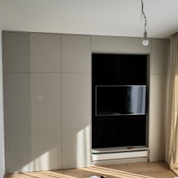 Eryk Pierzchała - Beżowa szafa wnękowa z miejscem na telewizor w salonie, z widocznymi zasłonami i elementami dekoracyjnymi. Minimalistyczny design.