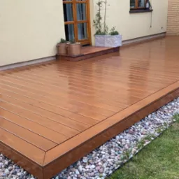 Świeżo polakierowany taras drewniany z deskami w odcieniach brązu, otoczony dekoracyjnymi kamieniami, przylegający do budynku z elementami zieleni.
