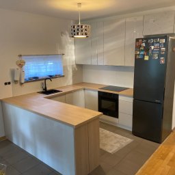 Eryk Pierzchała - Jasna kuchnia z drewnianym blatem, czarnym zlewem i lodówką ozdobioną magnesami. Nad blatem wiszą białe szafki, a nad nimi widoczne opakowania po sprzęcie.