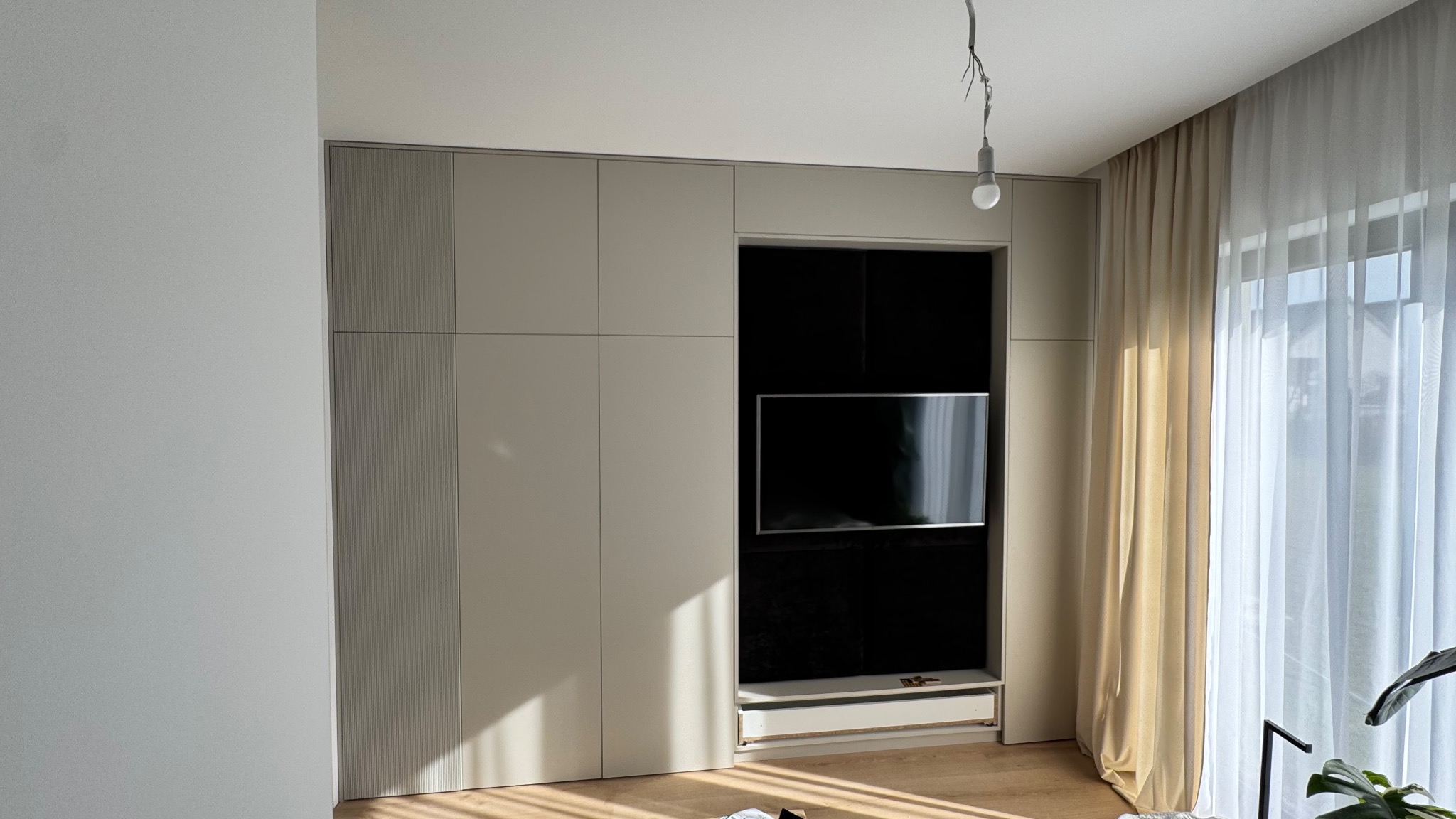 Beżowa szafa wnękowa z miejscem na telewizor w salonie, z widocznymi zasłonami i elementami dekoracyjnymi. Minimalistyczny design.