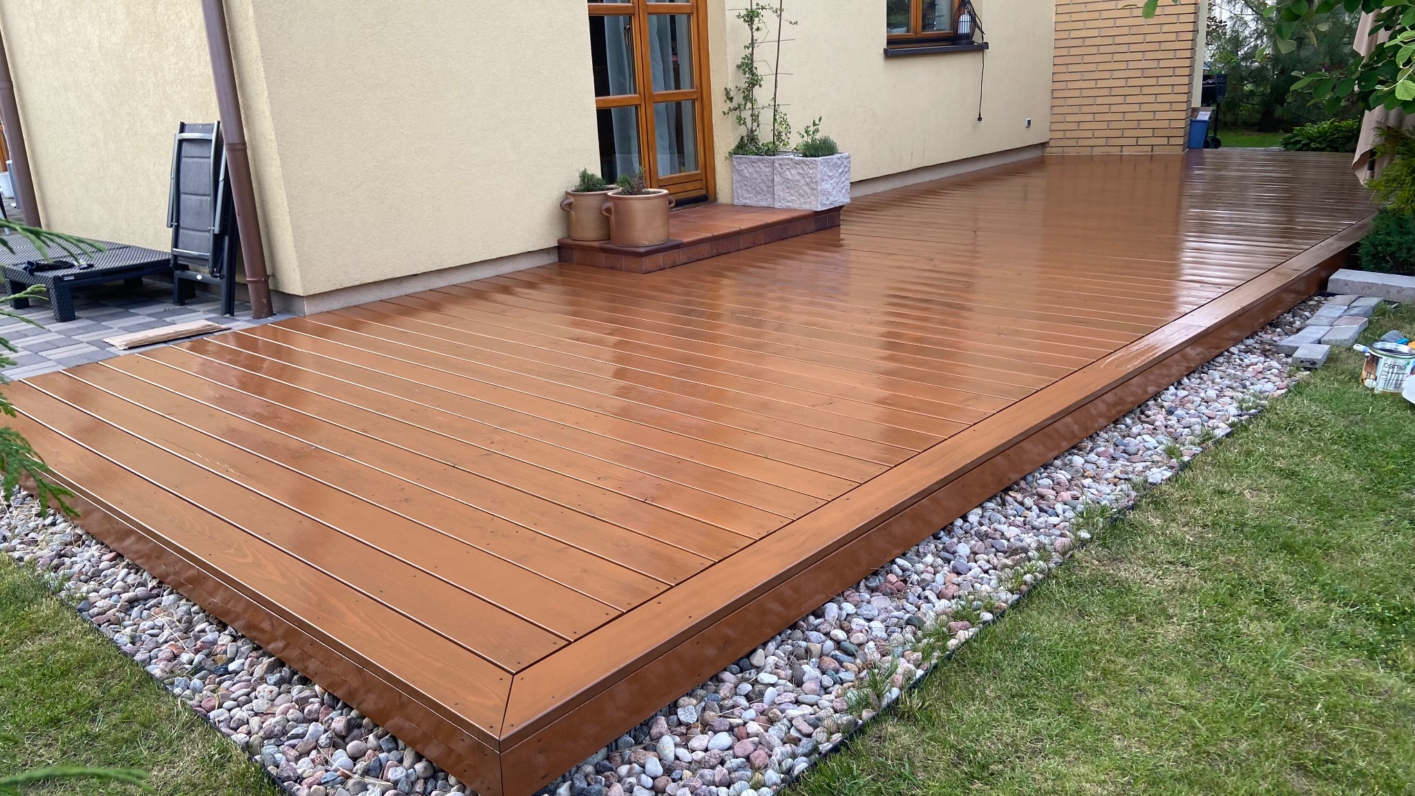 Świeżo polakierowany taras drewniany z deskami w odcieniach brązu, otoczony dekoracyjnymi kamieniami, przylegający do budynku z elementami zieleni.