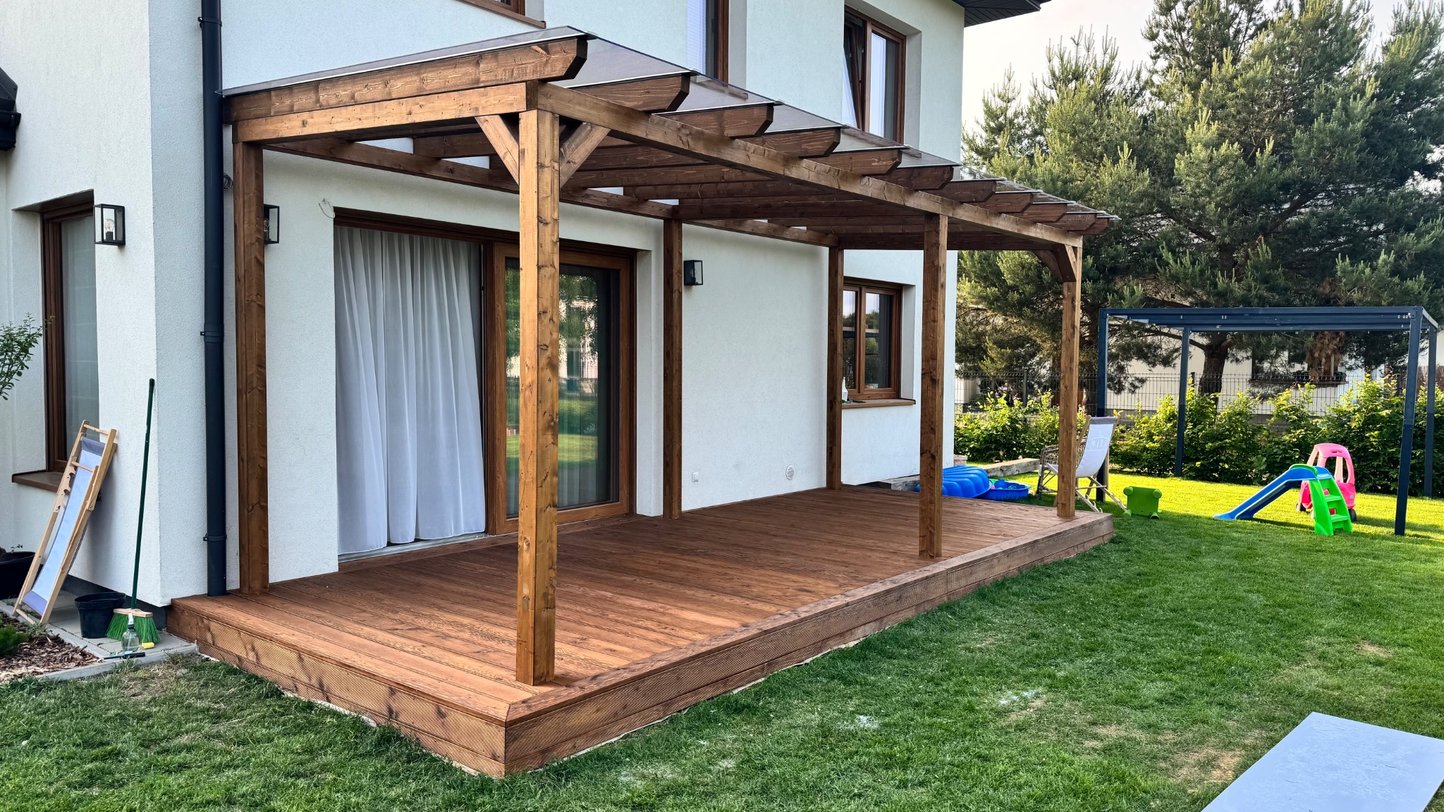 Drewniana pergola z przezroczystym zadaszeniem, przylegająca do domu z białymi ścianami. Widoczny taras z desek i zielony trawnik. W tle zabawki dziecięce.