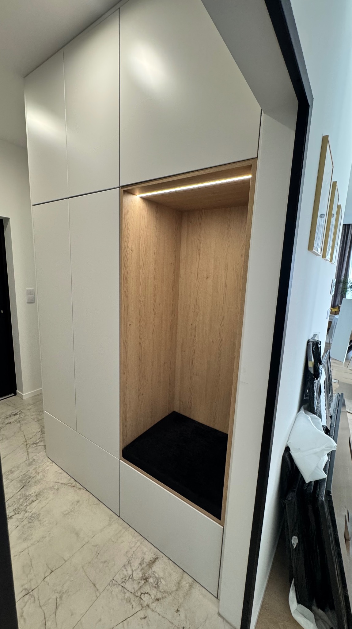Biała szafa wnękowa z drewnianą wnęką i czarnym siedziskiem, oświetlona listwą LED. Nowoczesny design, marmurowa podłoga, widok z perspektywy podłogi.