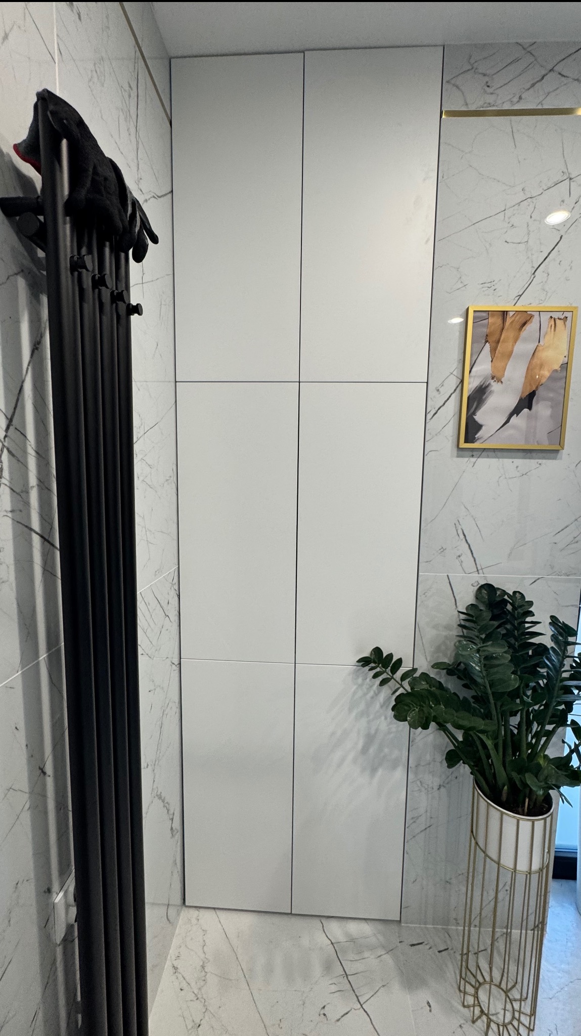 Biała szafa wnękowa z frontami bez uchwytów, na tle marmurowej ściany, z czarnym grzejnikiem żeberkowym i zieloną rośliną w ozdobnej donicy.