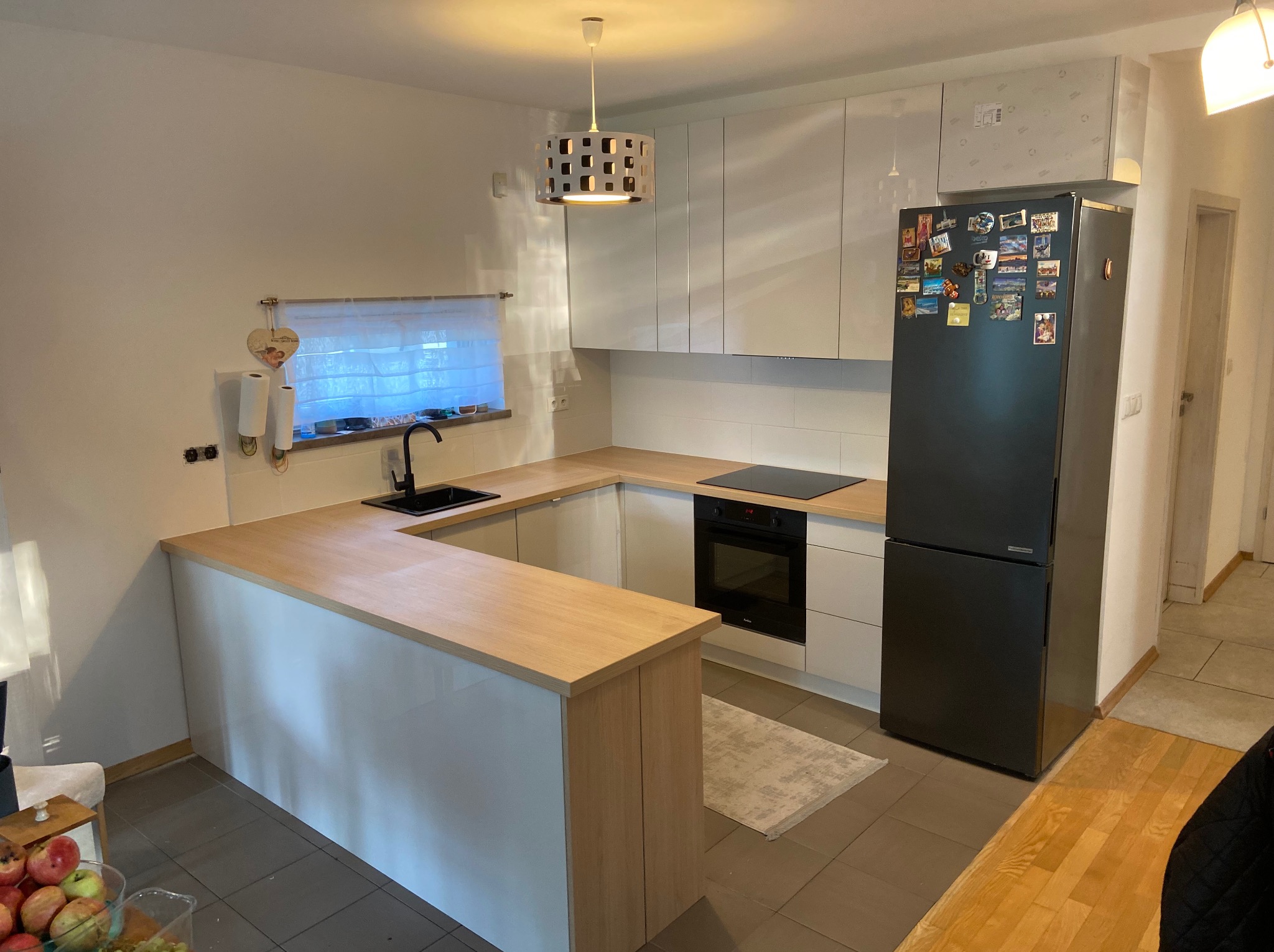 Jasna kuchnia z drewnianym blatem, czarnym zlewem i lodówką ozdobioną magnesami. Nad blatem wiszą białe szafki, a nad nimi widoczne opakowania po sprzęcie.