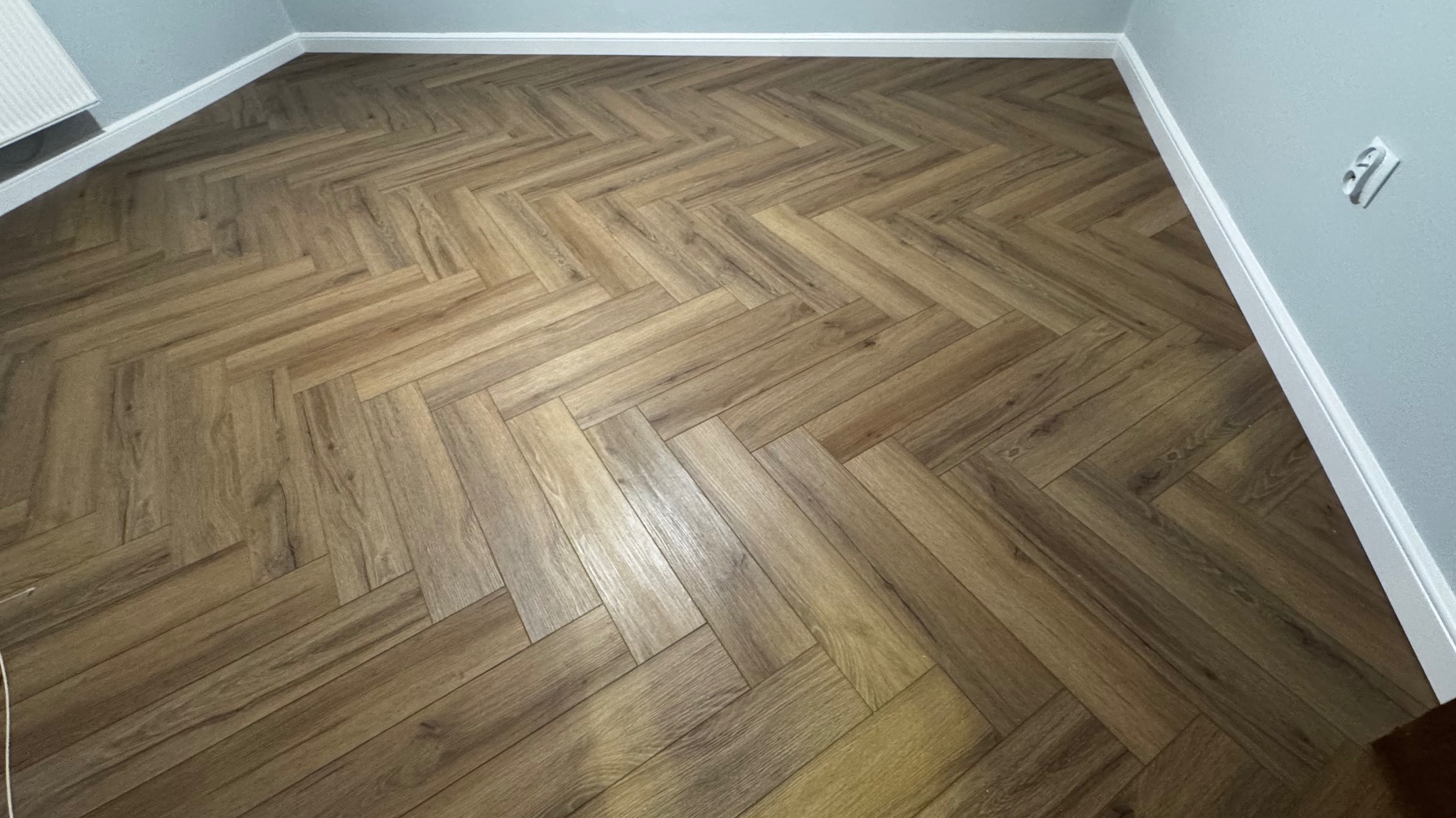 Ukończona podłoga z paneli ułożonych w jodełkę, z widocznymi listwami przypodłogowymi i gniazdkiem elektrycznym na ścianie. Ciepłe odcienie brązu i beżu.