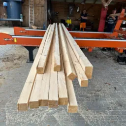 Świeżo przetarta tarcica z widocznymi śladami cięcia na tle tartaku z pracownikami, ułożona na pomarańczowej maszynie do obróbki drewna, ukazana w naturalnym oświetleniu.