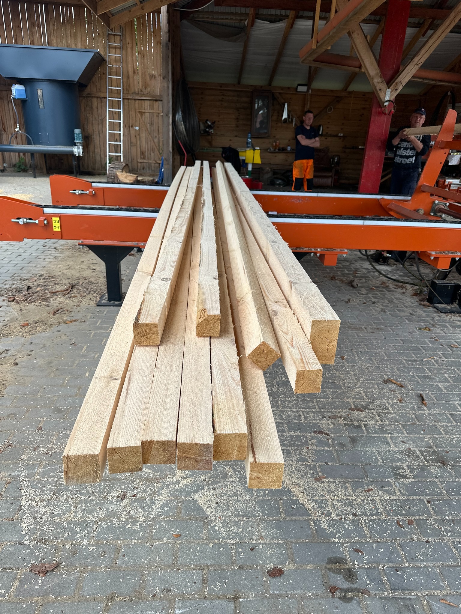 Świeżo przetarta tarcica z widocznymi śladami cięcia na tle tartaku z pracownikami, ułożona na pomarańczowej maszynie do obróbki drewna, ukazana w naturalnym oświetleniu.