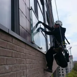 Pracownik w kasku, zawieszony na linach, czyści okna na wysokości na tle ceglanej elewacji i miejskiego krajobrazu z dźwigami budowlanymi.