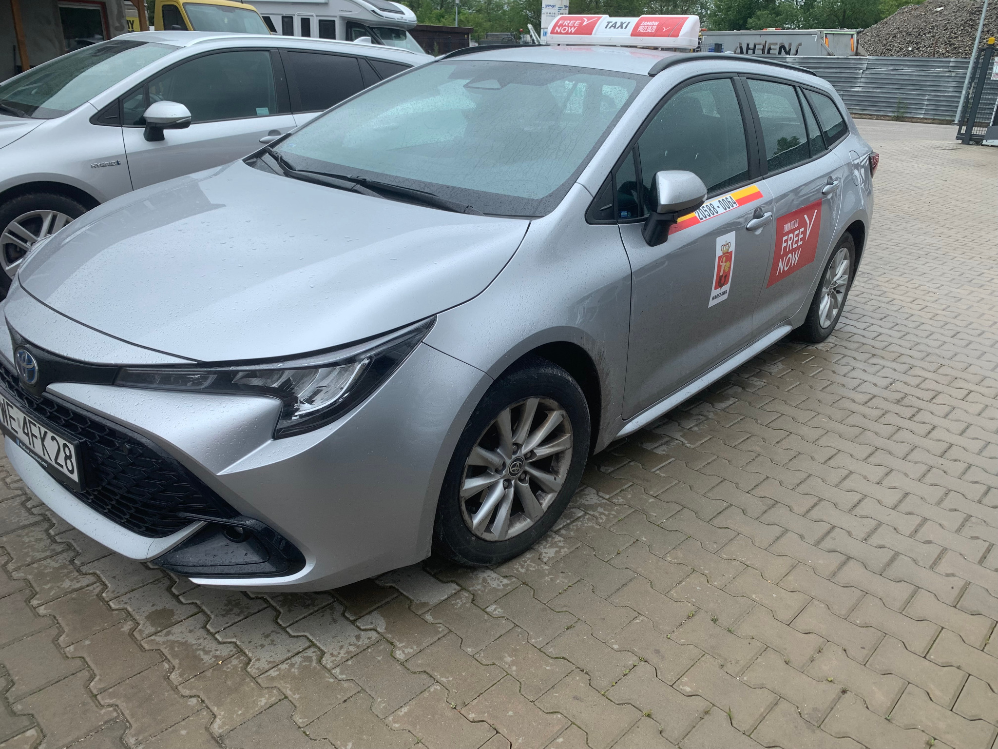 Srebrna Toyota Corolla kombi z oznaczeniami Taxi Free Now, zaparkowana na mokrym chodniku. Widoczne detale: logo, numer licencji, reklama na dachu.