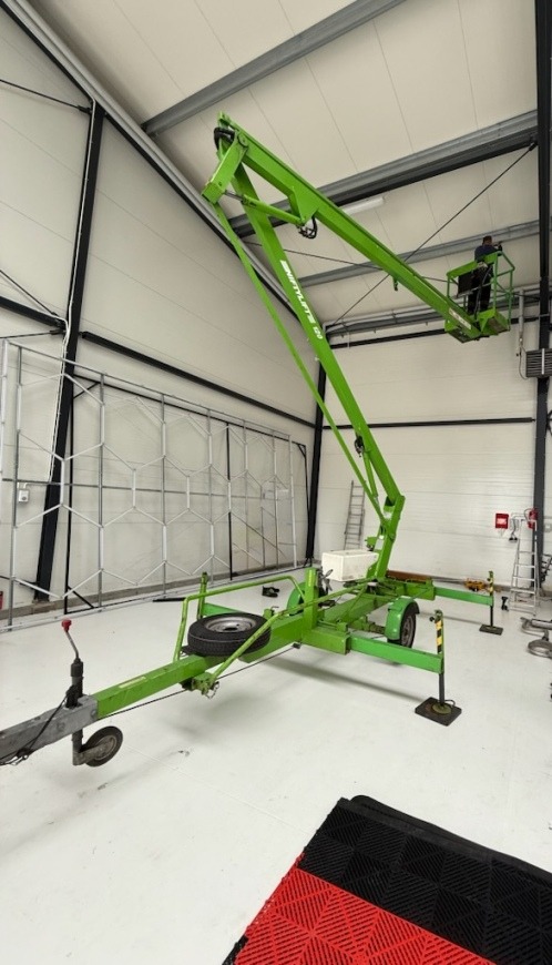 Zielony podnośnik koszowy Niftylift 120 w hali z pracownikiem w koszu, przygotowany do mycia elewacji lub dachu. Widoczna konstrukcja ściany.