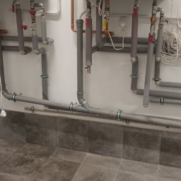 Oleksii Tkachov - Szare rury instalacji grzewczej/hydraulicznej przy ścianie, zabezpieczone izolacją. Widoczne zawory, złączki i uchwyty mocujące. Szara podłoga z płytek.