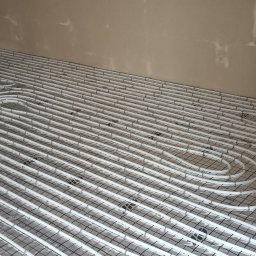 Oleksii Tkachov - Białe rury ogrzewania podłogowego ułożone na czarnej siatce, widoczne materiały budowlane w tle, ściany w trakcie wykańczania.