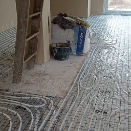 Oleksii Tkachov - Białe rury ogrzewania podłogowego na siatce, przygotowane do wylania posadzki w Toruniu. Widoczny fragment ściany i drabina malarska.