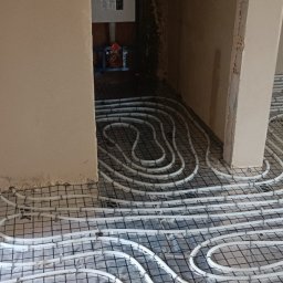 Oleksii Tkachov - Białe rury ogrzewania podłogowego ułożone na czarnej siatce w surowym wnętrzu, widok z góry, z fragmentami ścian w tle.