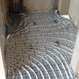 Białe rury ogrzewania podłogowego ułożone na czarnej macie z siatką, widoczne pionowe rury przy ścianie, surowy stan, instalacja przed wylaniem posadzki.