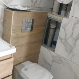 Oleksii Tkachov - Biała toaleta podwieszana z drewnianą obudową i chromowanym przyciskiem spłukującym, na tle marmurowych płytek. Widoczny bojler i instalacja.