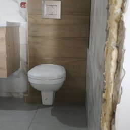 Oleksii Tkachov - Biała toaleta wisząca ze srebrnym przyciskiem spłukującym na tle drewnopodobnej zabudowy, szare płytki podłogowe i marmurowe płytki ścienne. Widoczna krawędź ściany w trakcie remontu.