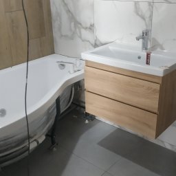 Oleksii Tkachov - Nowoczesna łazienka w trakcie wykończenia: wanna z hydromasażem i umywalka z drewnianą szafką na tle marmurowych płytek. Szare płytki podłogowe.