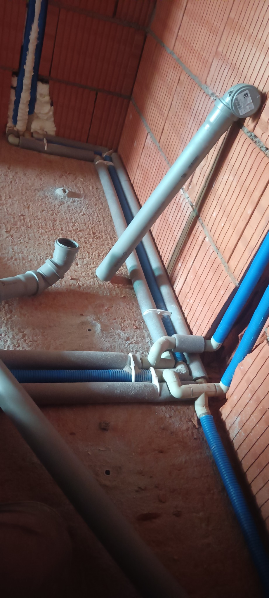 Szare i niebieskie rury instalacji hydraulicznej w surowym wnętrzu z ceglanymi ścianami. Rury częściowo zaizolowane, widoczne złącza i mocowania.