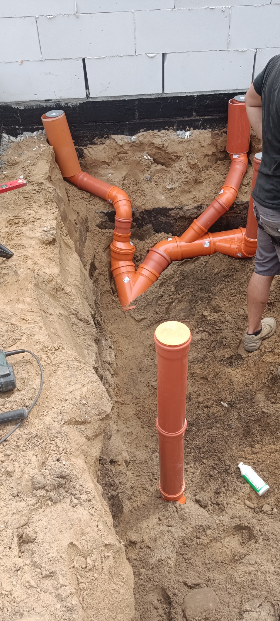 Pomarańczowe rury kanalizacyjne ułożone w wykopie ziemnym przy fundamencie budynku. Widoczny fragment instalacji i narzędzia pracy.