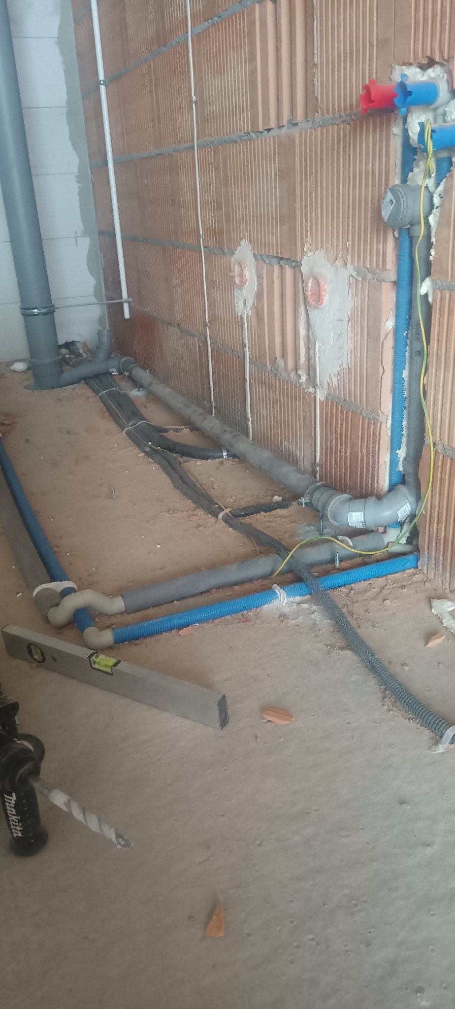 Instalacja wodno-kanalizacyjna w surowym wnętrzu: rury PCV, peszle i poziomica. Widoczne fragmenty ścian z cegły dziurawki i narzędzie.