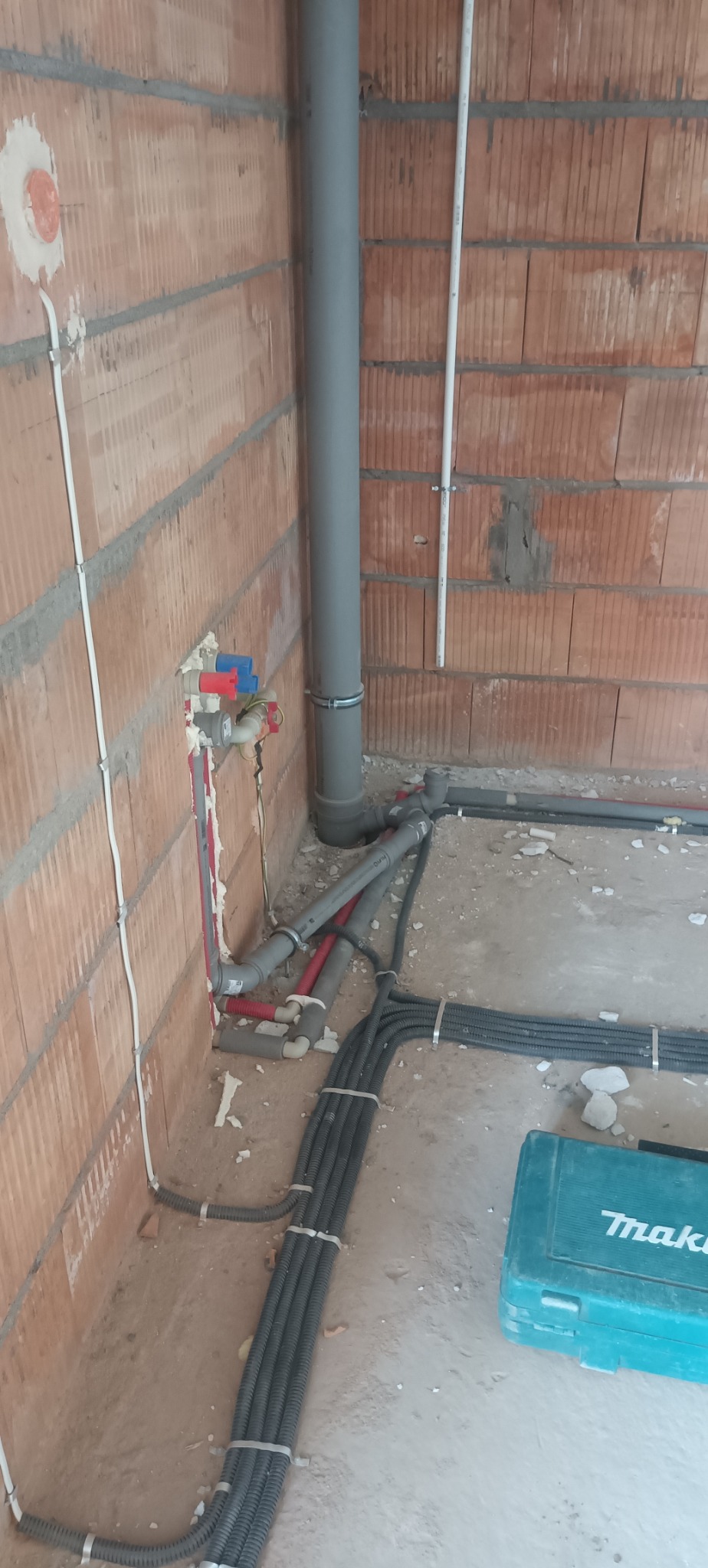 Surowa instalacja wodno-kanalizacyjna w trakcie montażu. Widoczne rury, zawory i kable na tle ceglanej ściany i surowej podłogi. W tle narzędzia w turkusowej skrzynce.