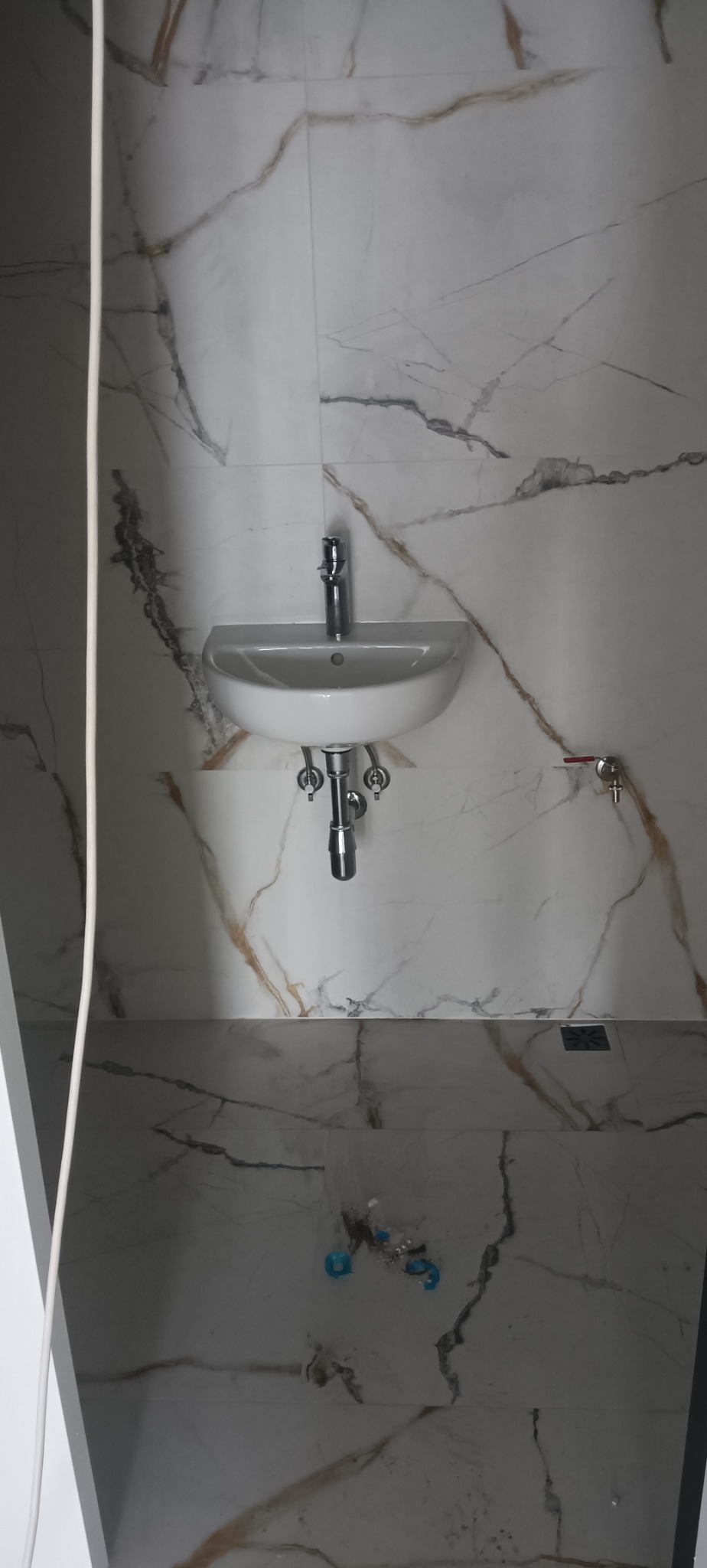 Biała, wisząca umywalka z chromowaną armaturą na tle płytek imitujących marmur z brązowymi żyłami. Widoczny zawór i fragment podłogi z odpływem.