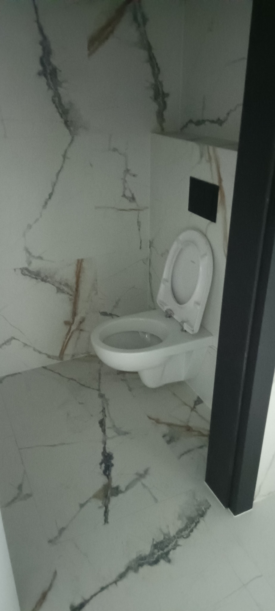 Biała toaleta wisząca w nowoczesnej łazience wykończonej płytkami imitującymi marmur z wyraźnym wzorem żyłkowania w odcieniach szarości i brązu. Widoczna czarna rama drzwi.