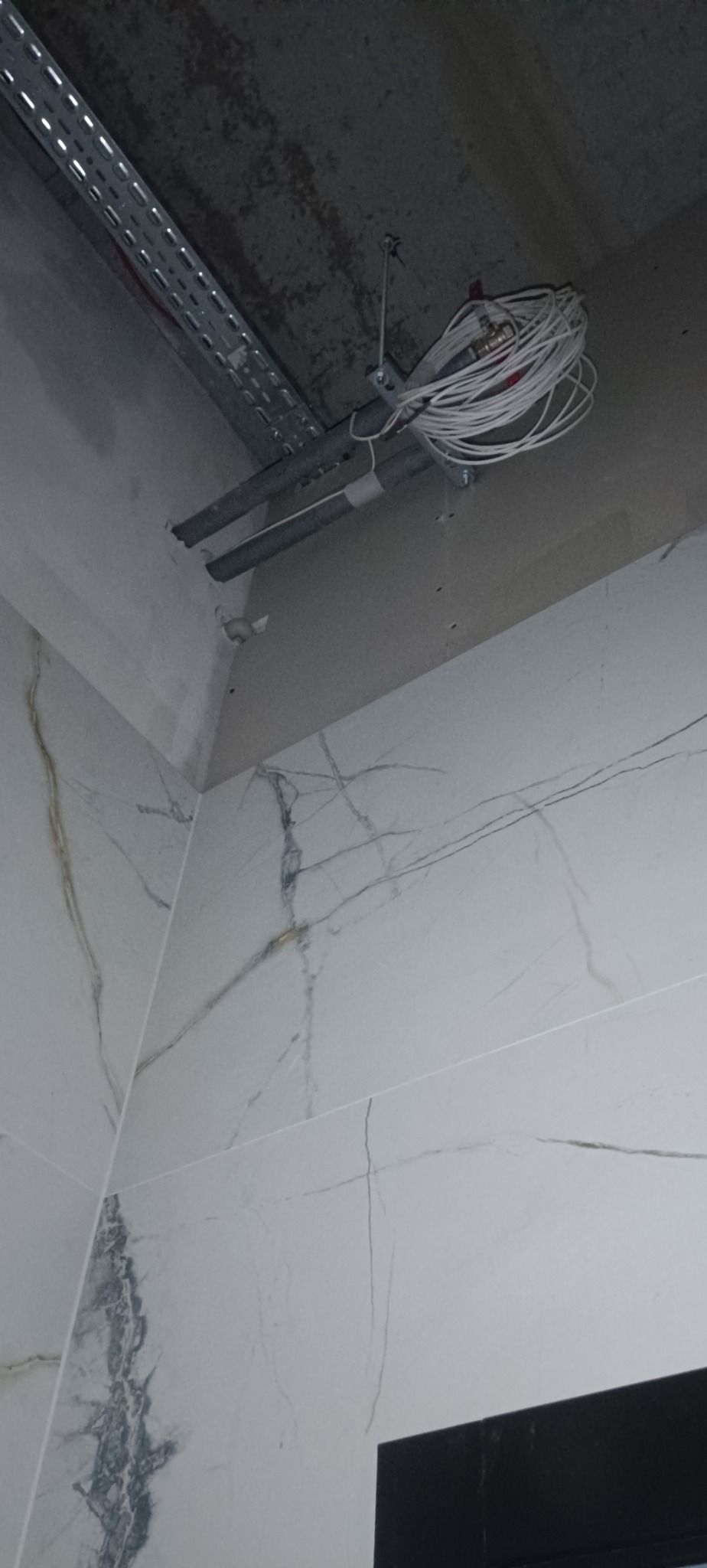 Wnętrze łazienki w trakcie prac: widoczne białe marmurowe płytki, rury instalacyjne pod sufitem oraz kable. Szyna montażowa pod sufitem.