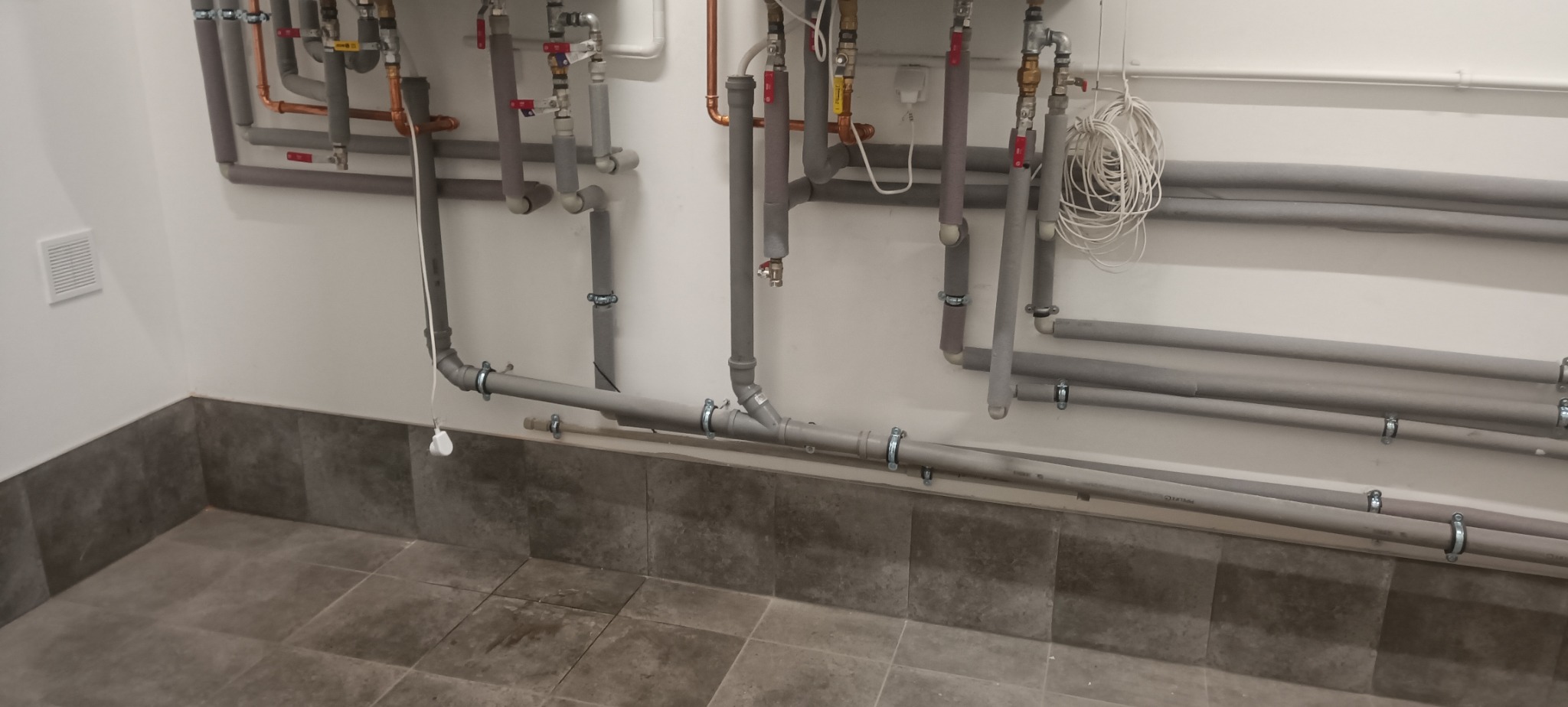 Szare rury instalacji grzewczej/hydraulicznej przy ścianie, zabezpieczone izolacją. Widoczne zawory, złączki i uchwyty mocujące. Szara podłoga z płytek.