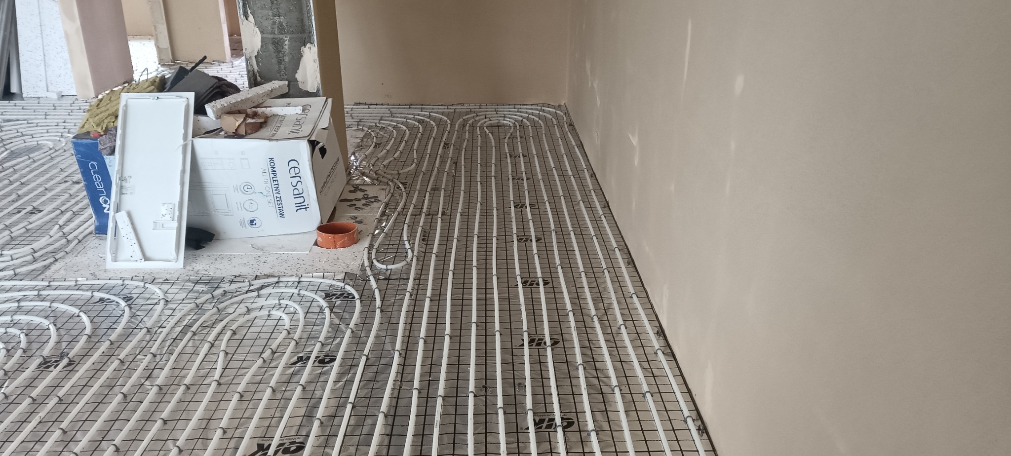 Białe rury ogrzewania podłogowego ułożone na siatce, widoczne pudełka z armaturą sanitarną. Etap instalacji w surowym wnętrzu.