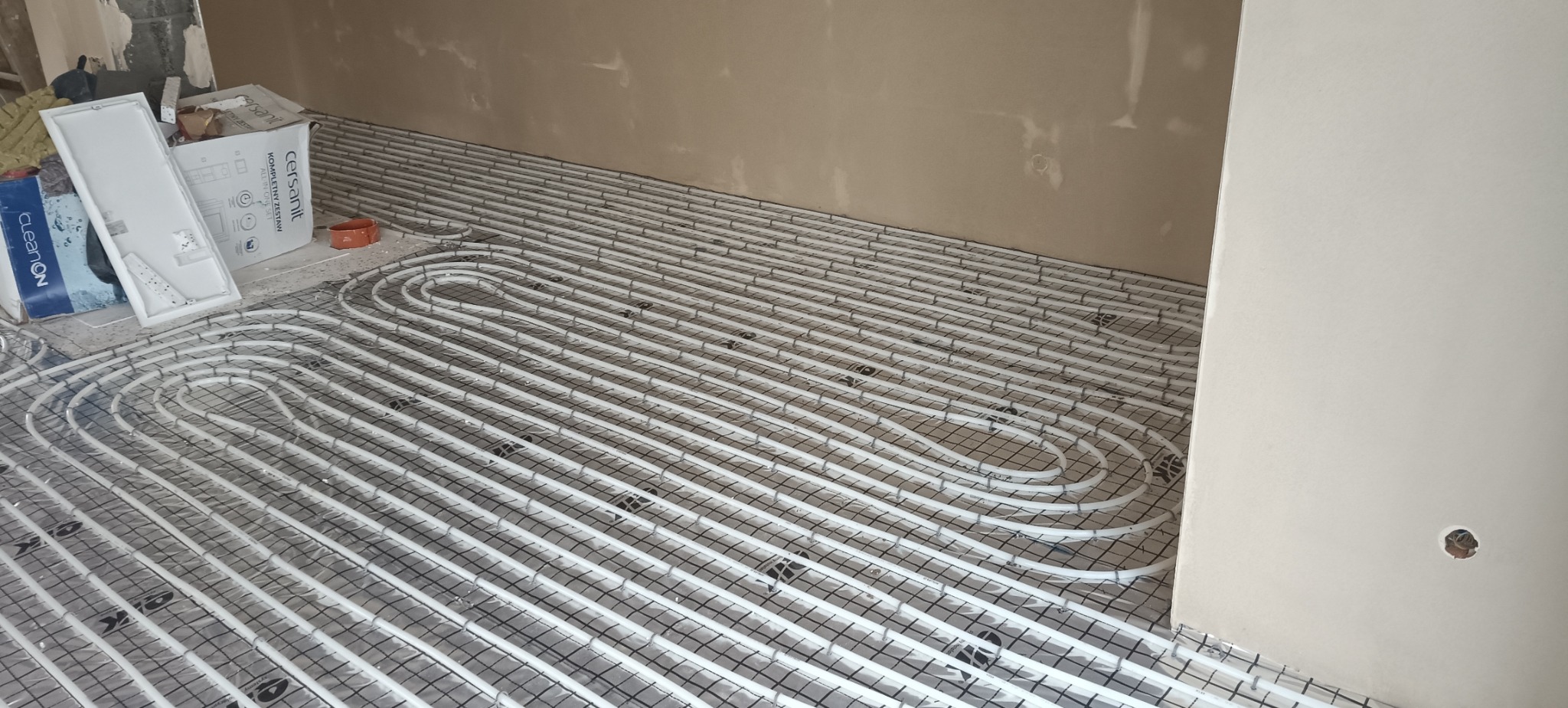 Białe rury ogrzewania podłogowego ułożone na czarnej siatce, widoczne materiały budowlane w tle, ściany w trakcie wykańczania.