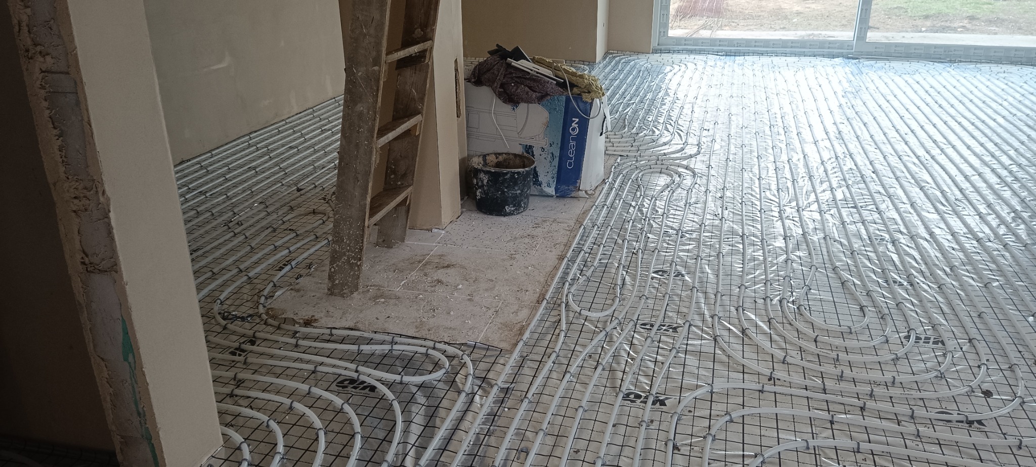 Białe rury ogrzewania podłogowego na siatce, przygotowane do wylania posadzki w Toruniu. Widoczny fragment ściany i drabina malarska.