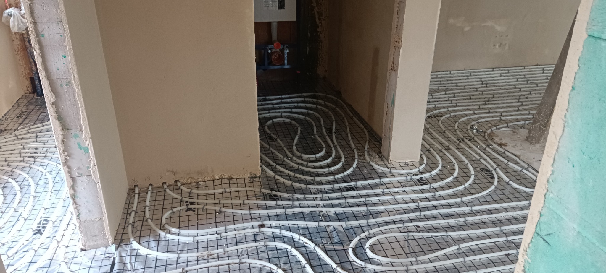 Białe rury ogrzewania podłogowego ułożone na czarnej siatce w surowym wnętrzu, widok z góry, z fragmentami ścian w tle.