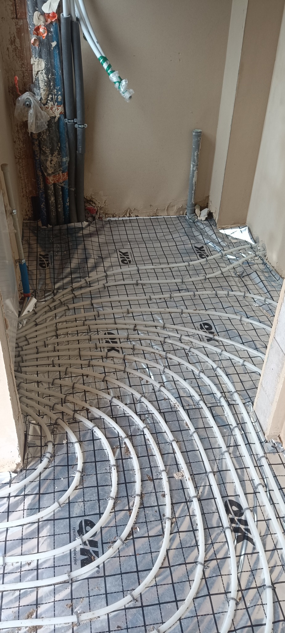 Białe rury ogrzewania podłogowego ułożone na czarnej macie z siatką, widoczne pionowe rury przy ścianie, surowy stan, instalacja przed wylaniem posadzki.