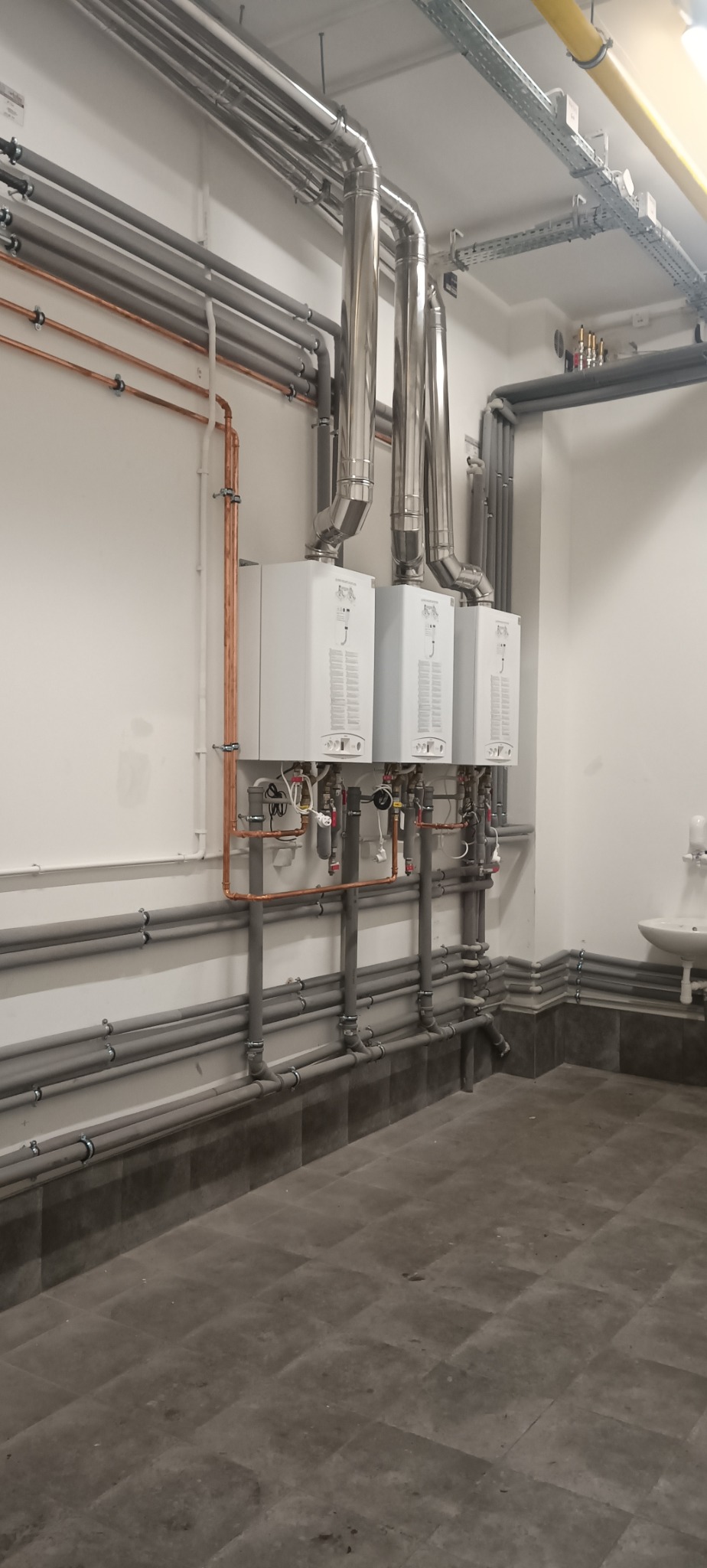 Trzy białe piece gazowe z rurami miedzianymi i stalowymi na ścianie. Szare rury i płytki podłogowe. Widok na instalację grzewczą w pomieszczeniu.