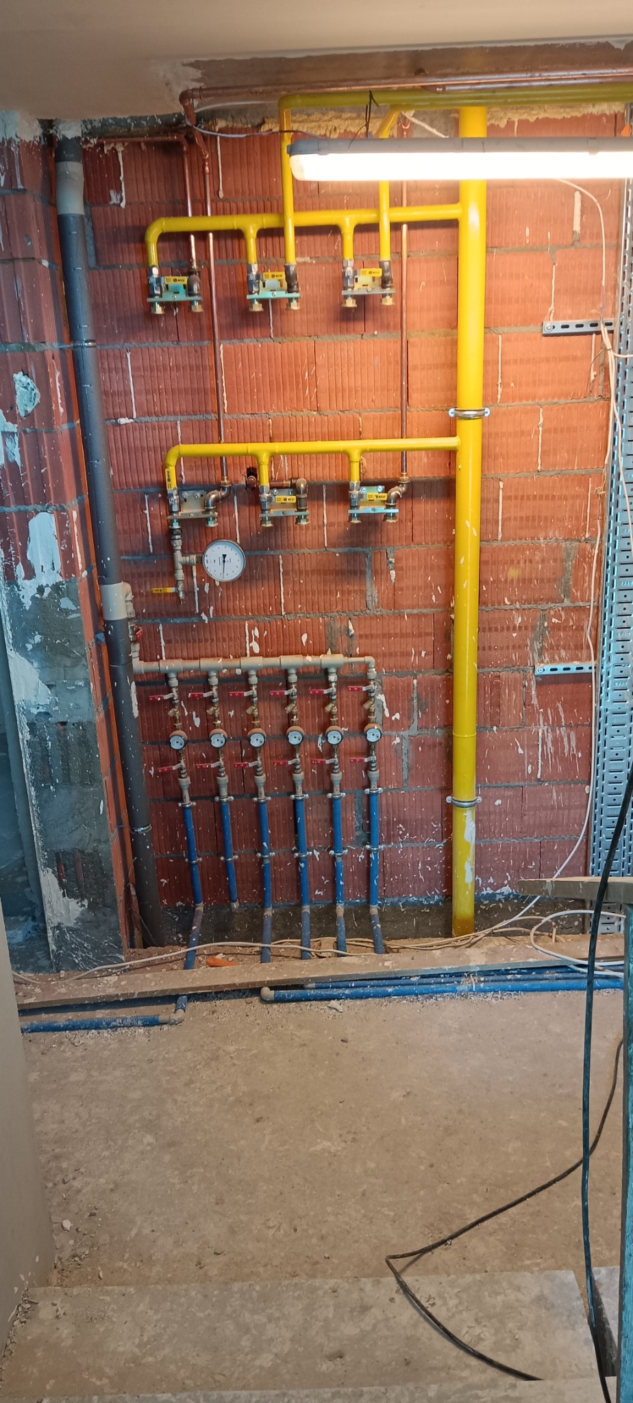 Instalacja gazowa i hydrauliczna z żółtymi rurami na tle ceglanej ściany. Widoczne zawory, manometr i przewody. Prace instalacyjne w surowym wnętrzu.