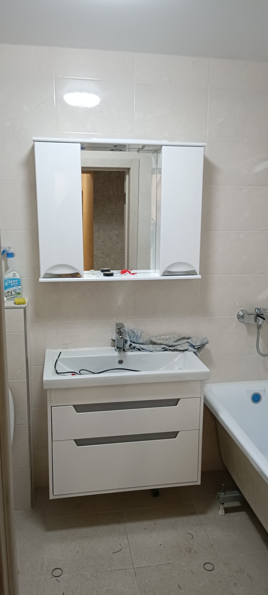 Biała umywalka z szafką i lustrem w łazience wyłożonej jasnymi płytkami. Widoczna wanna i akcesoria łazienkowe.