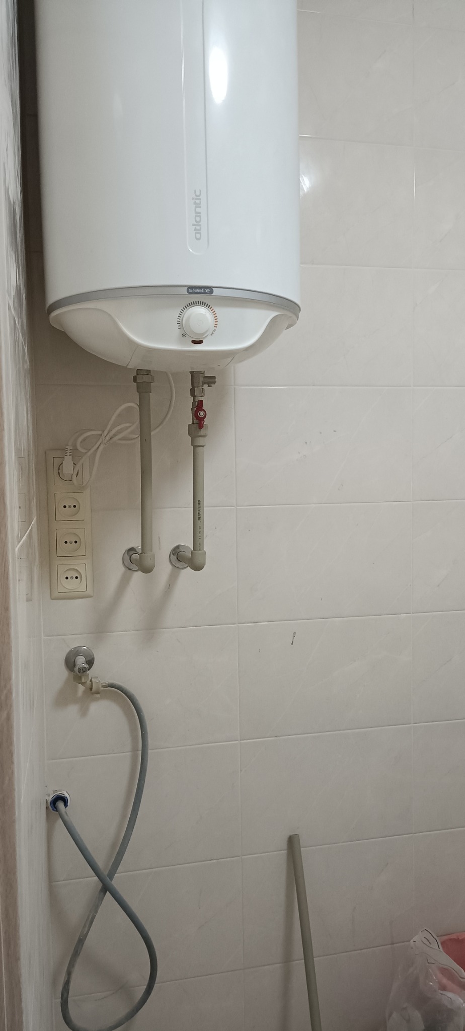 Biały, wiszący bojler Atlantic z podłączonymi rurami na tle jasnych, kwadratowych płytek. Widoczne gniazdka elektryczne i szara rura doprowadzająca wodę.